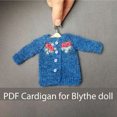 a hand holding a small blue knitted sweater with flowers on it and the words pdf cardigan for blythe doll