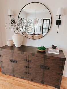 a large round mirror hangs on the wall above a dresser with drawers and vases