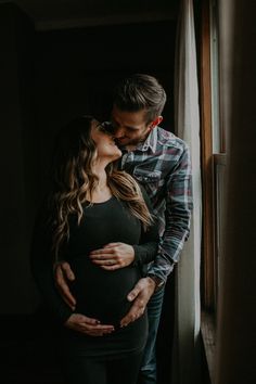 a pregnant couple kissing while standing in front of a window