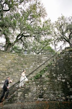 a man and woman walking up the stairs in front of a stone wall with trees