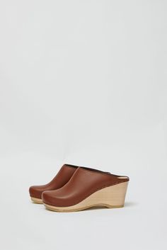 Sleek, closed-toe clog slide on wedge heel. British tan leather upper. Rounded toe, open back, and minimal stitching. Lightweight, lacquered wood base. Unlined. Every clog is handmade. Textures of leather can vary from shoe to shoe. Certain styles may take up to 10 business days to arrive. Modern Brown Slip-on Clogs, Modern Clogs With Wooden Wedge Heel, Brown Wedge Heel Mules With Rubber Sole, Brown Wedge Heel Clogs With Rubber Sole, Brown Mules With Rubber Sole And Wedge Heel, Brown Clogs With Rubber Sole And Wedge Heel, Brown Wedge Heel Clogs With Leather Sole, Brown Leather Sole Wedge Clogs, Brown Wedge Heel Mules With Deep Heel Cup