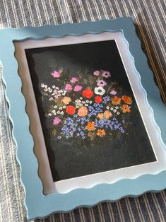 a blue frame with flowers painted on it sitting on a striped table cloth next to a vase