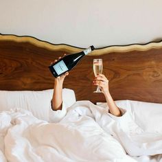 a woman laying in bed with a glass of wine and bottle on her head while holding a champagne flute