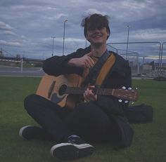 a man sitting on the grass holding a guitar in his hands and smiling at the camera