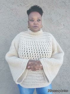 a woman standing in front of a gray wall wearing a white knitted sweater and blue jeans