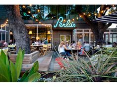 people sitting at tables in front of a restaurant with lights strung from the roof and trees