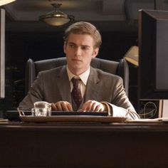 a man in a suit sitting at a desk with a computer and cup on it