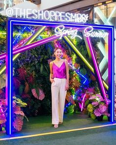 a woman standing in front of a neon sign with flowers and plants on it's sides