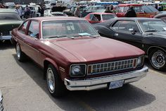 an old red car parked in a parking lot next to other older cars and people