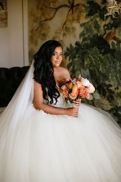 a woman in a wedding dress sitting on a couch holding a bridal bouquet and looking at the camera