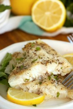 a white plate topped with fish next to lemon wedges and a fork on top of a wooden table