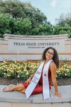 a woman sitting on the side of a cement wall wearing a sash and smiling at the camera