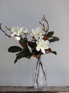 white flowers in a clear glass vase on a wooden table with grey wall behind it