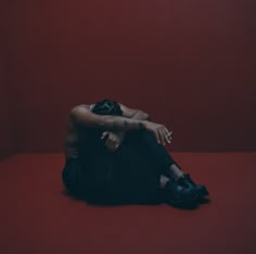 a woman sitting on the floor with her arms crossed and hands behind her head, in front of a red wall