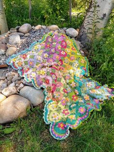 a crocheted blanket is laying on the ground next to some rocks and trees