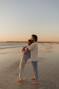 a man and woman standing on top of a beach next to the ocean at sunset