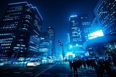 people are standing on the sidewalk in front of some tall buildings at night with bright lights