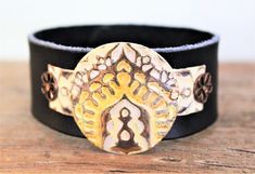 I created this one-of-a-kind genuine metal and leather cuff bracelet featuring a decorative design on natural brass blanks that have been riveted and attached to the leather bracelet with decorative washers and rivets. A mix of cream and yellow patina paints complete this piece. The leather band is colored with lead-free vegetable dyes. Your bracelet will arrive packaged and ready for gift giving.  The bracelet fastens in two different locations and is adjustable as follows: 1st Snap @ approx. 7 inches (inside measurement) 2nd Snap @ approx. 8 inches (inside measurement) Total Bracelet Length (end to end): 9 inches Width: 1 inch Depth: 1.5mm Care Instructions: DO NOT exercise/work out, shower, bathe or swim while wearing this bracelet; it is not waterproof. Avoid contact with hairsprays, p Unique Leather Cuff Bracelet For Festival, Adjustable Traditional Leather Bracelet, Bohemian Stamped Leather Bracelet, Adjustable Stamped Leather Cuff Bracelet, Artisan Adjustable Cuff Leather Bracelet, Adjustable Leather Strap Bohemian Cuff Bracelet, Bohemian Cuff Leather Bracelet Gift, Adjustable Bohemian Cuff Bracelet With Leather Strap, Traditional Adjustable Cuff Bracelet With Concho