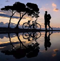 two people are standing next to their bikes on the water at sunset or dawn with trees and clouds reflected in the water