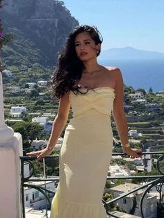 a woman in a white dress standing on a balcony overlooking the ocean and mountains with her arms out