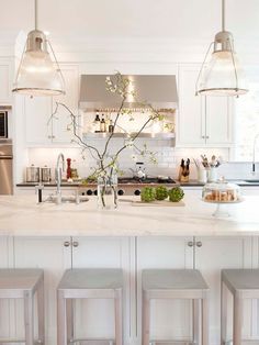 a white kitchen with four stools in front of the island and three lights hanging from the ceiling