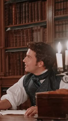 a man sitting at a table in front of a book shelf with candles on it