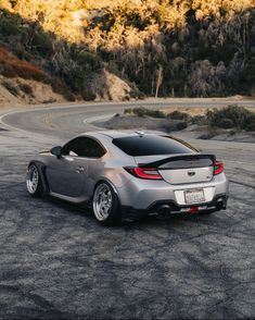 the rear end of a silver sports car on a road with mountains in the background