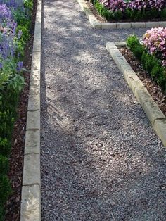 a gravel path lined with purple flowers and greenery next to a sidewalk in the middle of a garden