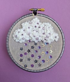 an embroidery project with white flowers and purple stars on a gray hoop hanging on a purple wall