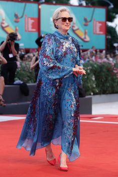 an older woman in a blue dress walking on a red carpet with people behind her