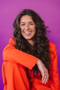 a smiling woman in an orange outfit with her arms on her chest, sitting against a purple background