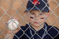 a young boy holding a baseball behind a chain link fence with his face painted like a bat