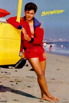 a man in red swimsuit standing on beach next to yellow surfboard with words baywatch