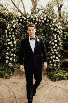 a man in a tuxedo is walking towards the camera with white flowers behind him