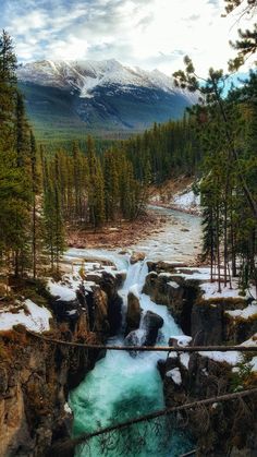 a river running through a forest filled with trees and snow covered mountains in the background