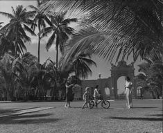 black and white photograph of people on bicycles in front of palm trees with an old building