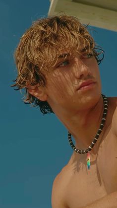 a shirtless young man holding a surfboard under his arm and wearing a beaded necklace