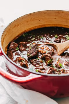 a red pot filled with beef stew on top of a white cloth next to a wooden spoon