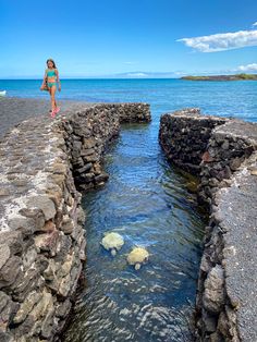 a woman walking along the edge of a stone wall next to the ocean with rocks in the water