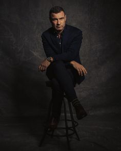 a man sitting on top of a stool in front of a black background wearing a suit