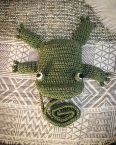 a crocheted green frog laying on top of a bed next to a pillow