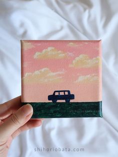 a hand holding up a small painting with a van in the sky on it's side