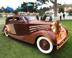 an antique car is parked on the grass in front of a white fence and some people