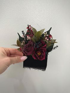 a hand holding a black and red flower arrangement in it's left side, against a white wall