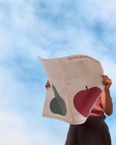 a woman holding up a newspaper with an apple and pear design on it, in front of a blue sky