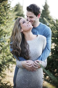 a man and woman standing next to each other in front of christmas tree trees with their arms around each other