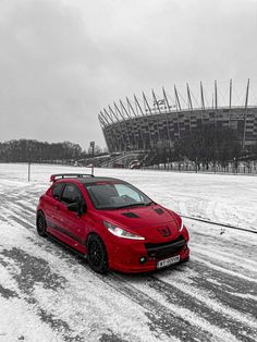 a red car driving down a snowy road next to a large stadium in the background