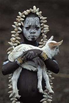 a woman with white face paint holding a baby goat in her arms and headdress