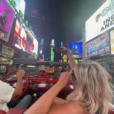 two people in a red convertible car driving through times square at night with neon signs and billboards