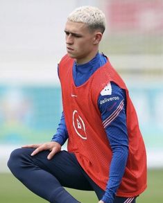 a young man sitting on top of a soccer field wearing a red shirt and blue pants